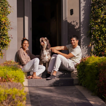 Two people sitting on a porch with a dog, surrounded by greenery and a house in the background.