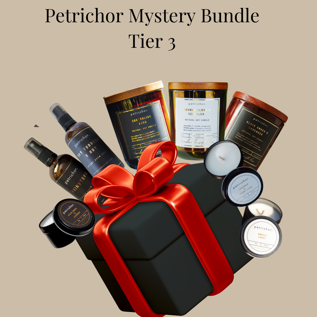 A Petrichor Mystery Bundle Tier 3 gift set featuring a black gift box with a red ribbon, surrounded by various home fragrance products including candles, diffusers, and a spray bottle, on a solid beige background.
