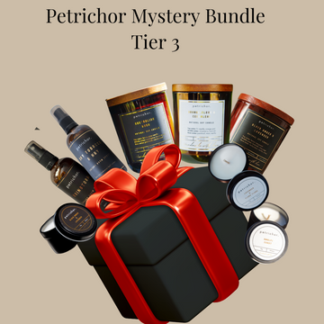 A Petrichor Mystery Bundle Tier 3 gift set featuring a black gift box with a red ribbon, surrounded by various home fragrance products including candles, diffusers, and a spray bottle, on a solid beige background.