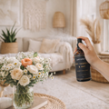 Hand holding a bottle of petrichor dry tobacco & hay room spray in a living room setting with flowers and decor.