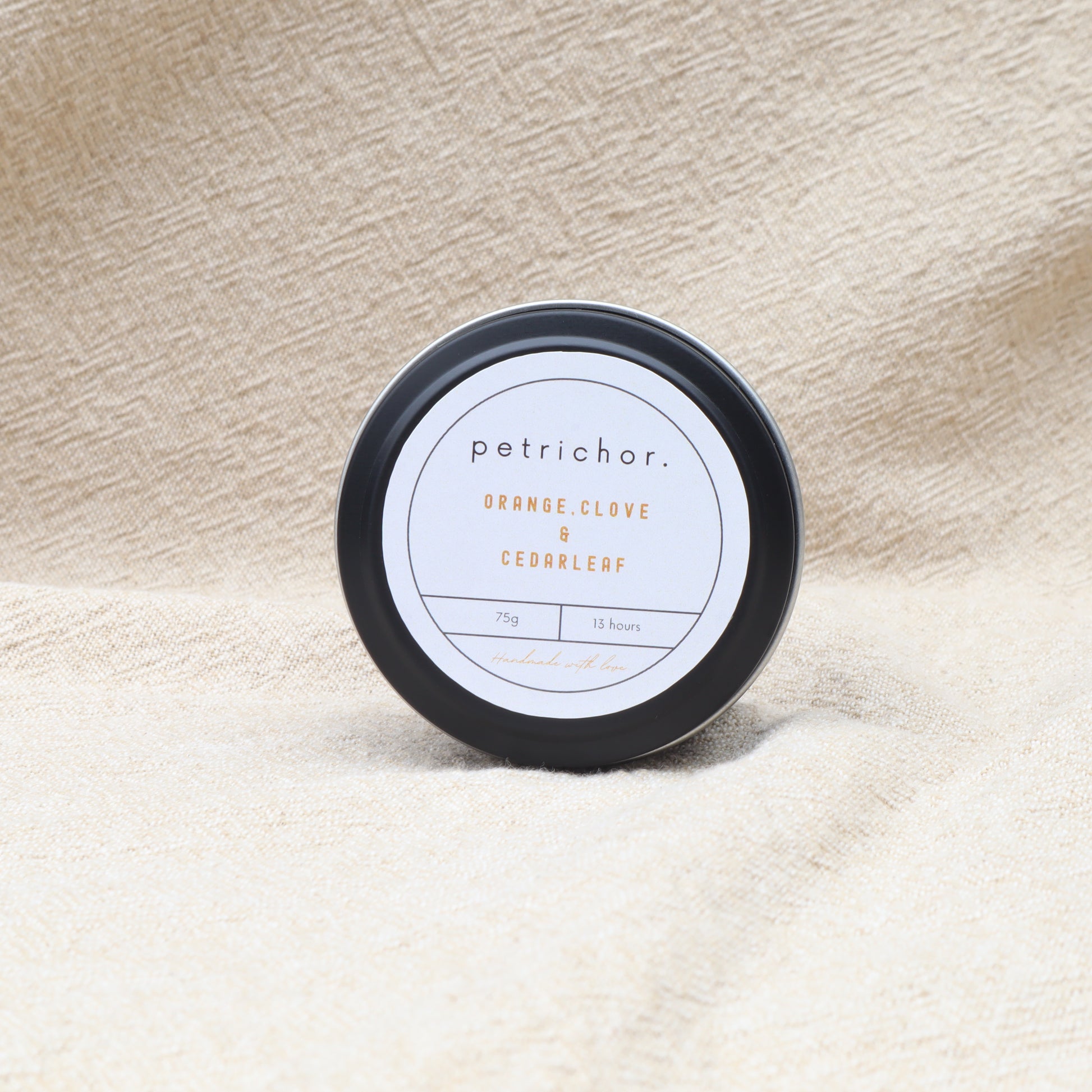 Candle tin labeled 'Petrichor' with 'Orange Clove & Cedarleaf' on a textured beige background
