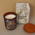 Candle in a decorative tin with a book titled 'Postcards from Spain' on a beige surface.