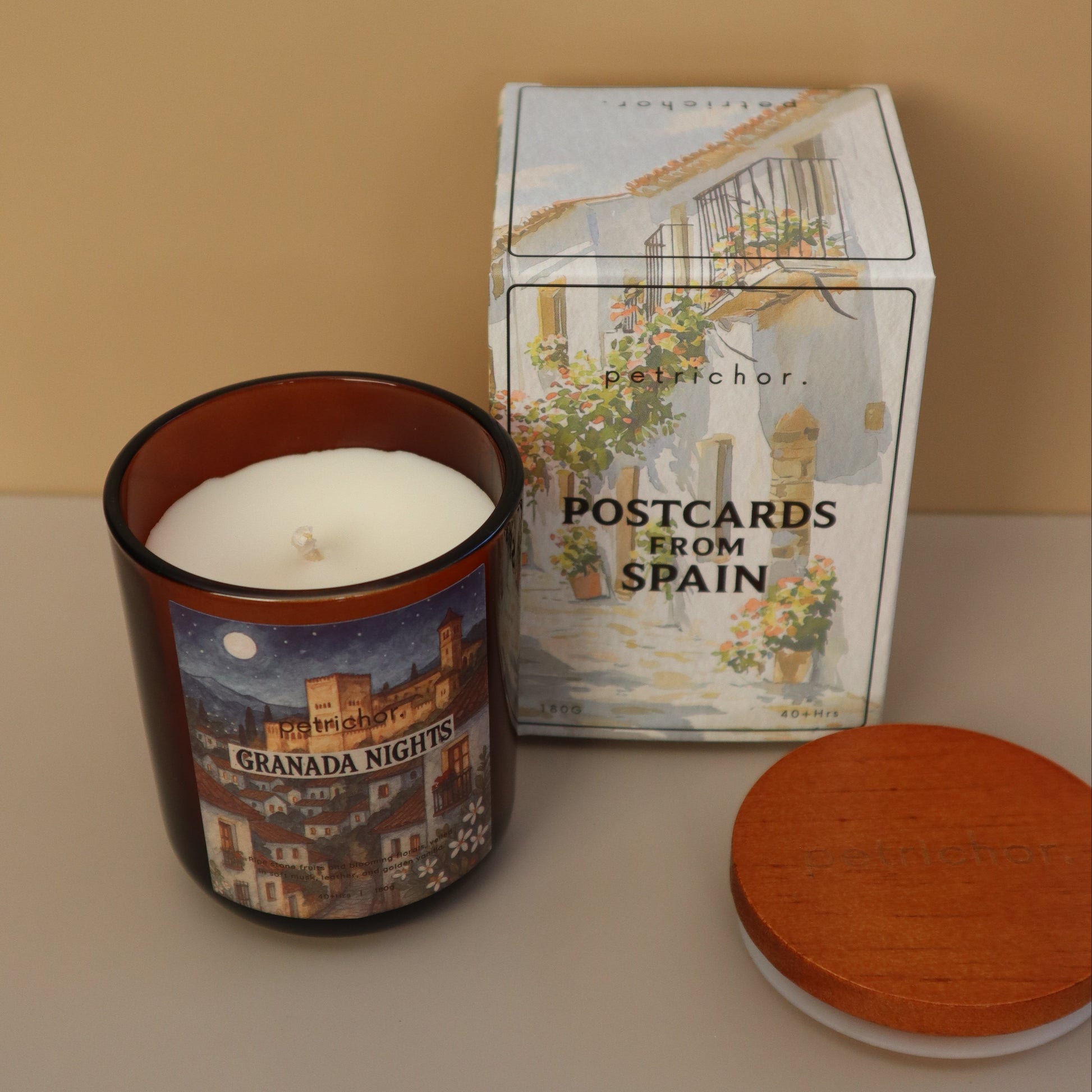 Candle in a decorative tin with a book titled 'Postcards from Spain' on a beige surface.