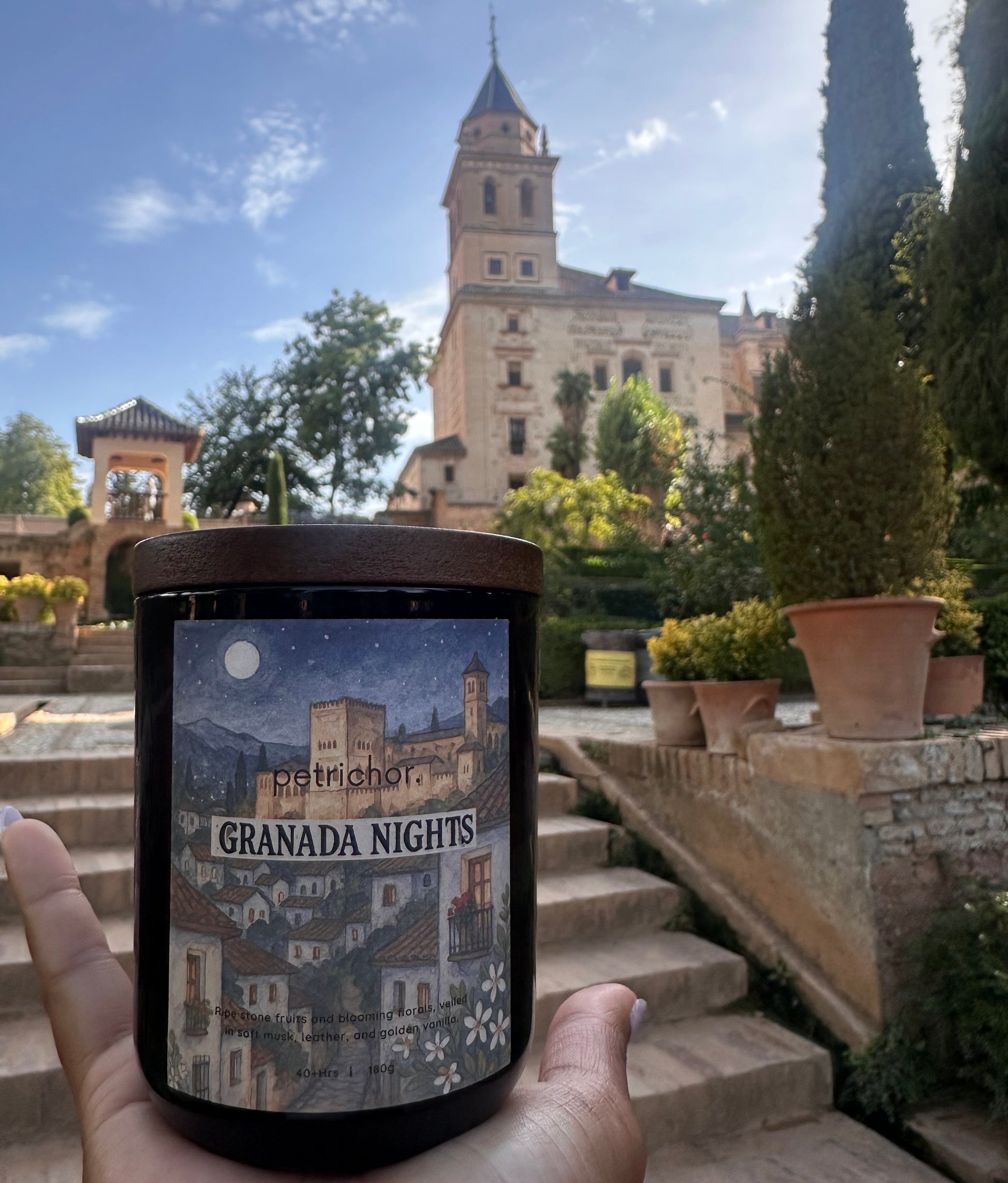 Hand holding a smartphone with a Granada Nights candle in front of a scenic view with a tower and blue sky.