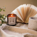 Candle with a book and mug on a soft surface, surrounded by plants