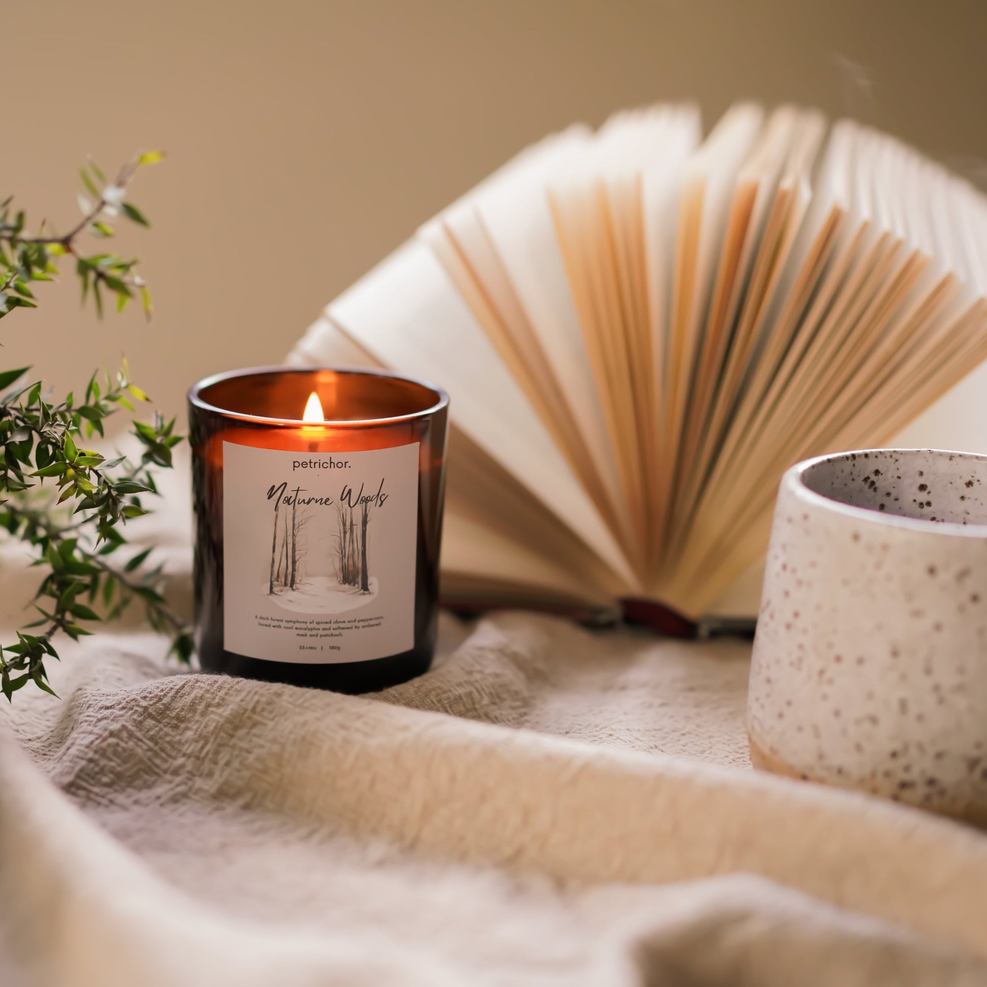 Candle with a book and mug on a soft surface, surrounded by plants